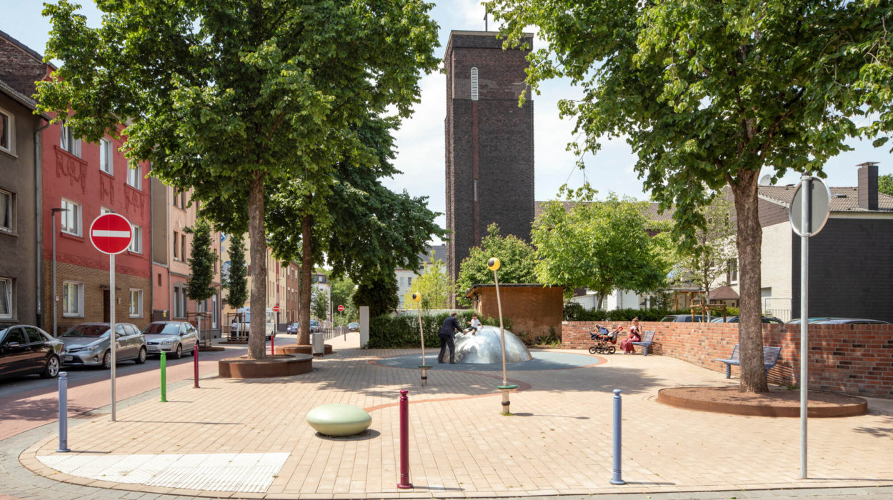 Man sieht eine Spielstraße mit einer Kuppel aus Metall auf der ein kleiner Junge rutscht. Eine männliche Person steht davor und fängt den Jungen auf. An der Straße sind Autos geparkt und es ist zu erkennen,dass es sich um eine Einbahnstraße handelt, da ein Einfahrt Verboten Schild an der Straßenecke steht.